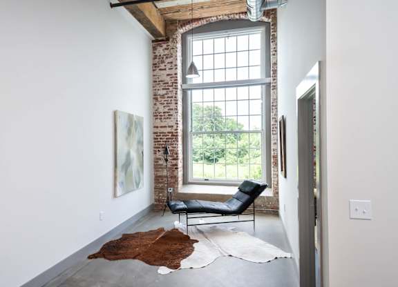 Loft Apartments Near Downtown Spartanburg, SC - Converse Mill Lofts - Living Room with High Ceiling, Large Window, Chaise Lounge, Pendant Lighting, Couch, Coffee Table, Armchairs, and Paintings.