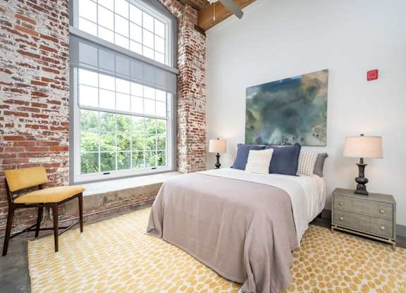 Loft Studios Spartanburg, SC - Converse Mill Lofts - Bedroom with High Ceiling, Concrete Flooring, Large Bed, Large Painting, Nightstands, Lamps, Chair, Large Windows, and Exposed Brick Walls.