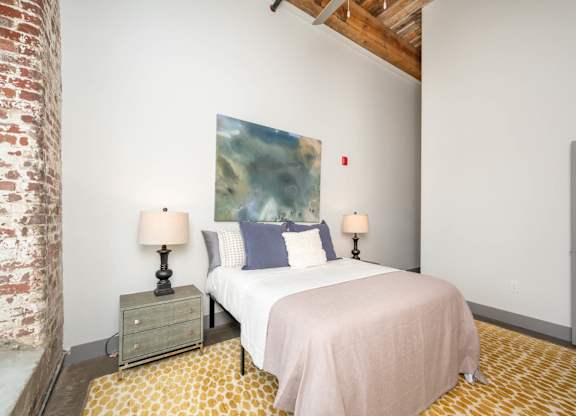 Loft Studios Spartanburg, SC - Converse Mill Lofts - Bedroom with High Ceiling, Concrete Flooring, Large Bed, Large Painting, Nightstands, Lamps, Chair, Large Windows, and Exposed Brick Walls.