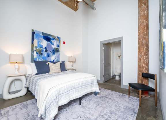 Loft Studios Spartanburg, SC - Converse Mill Lofts - Bedroom with High Ceiling, Concrete Flooring, Large Bed, Large Painting, Nightstands, Lamps, Chair, Large Windows, and Exposed Brick Walls.