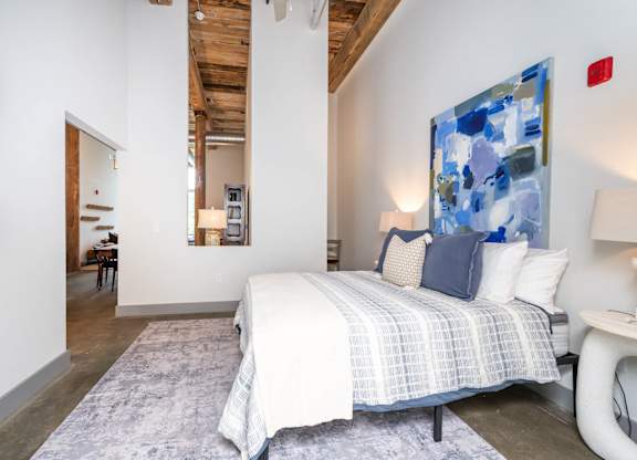 Loft Studios Spartanburg, SC - Converse Mill Lofts - Bedroom with High Ceiling, Concrete Flooring, Large Bed, Large Painting, Nightstands, Lamps, Chair, Large Windows, and Exposed Brick Walls.