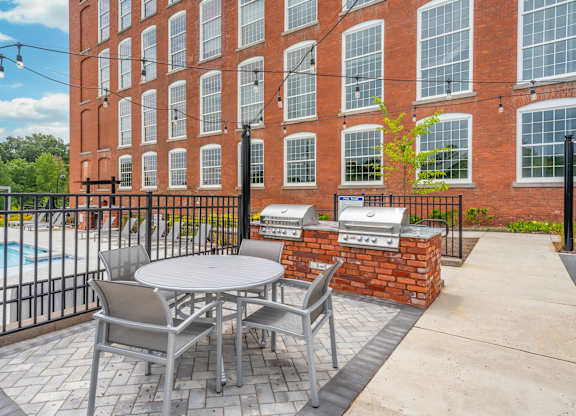 Loft Studio Apartments Spartanburg, SC - Converse Mill Lofts - Grilling Station with Brick Base, Built-In Stainless Steel Grills, Round Dining Table, Chairs, Black Metal Fence, and String Lights.