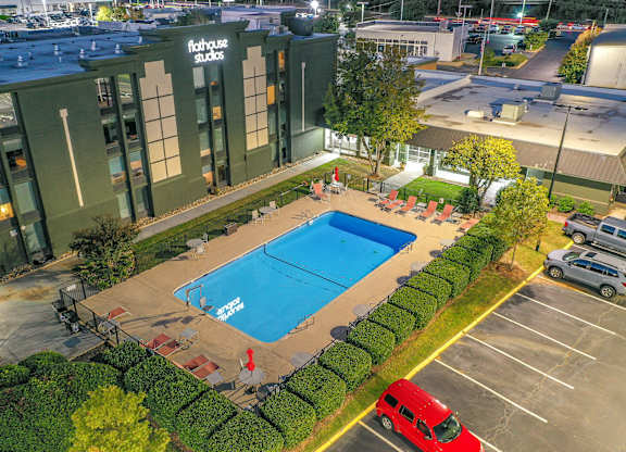 A swimming pool is surrounded by a hedge and a red van is parked in the parking lot.