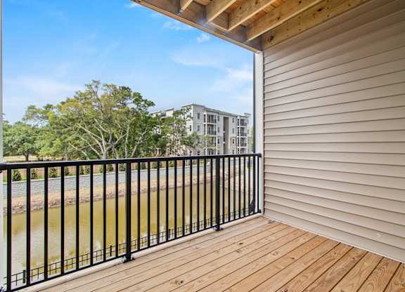 A balcony with a black railing and wooden floor overlooks a body of water and buildings.