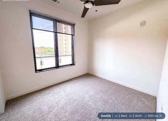 A second bedroom with a carpeted floor, a ceiling fan, and a large window overlooking a cityscape.