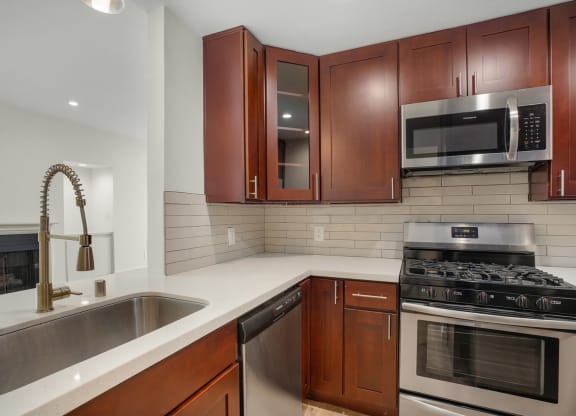 Apartment kitchen with stainless steel appliances, white tile back splash, white counter tops, and dark wood cabinets.