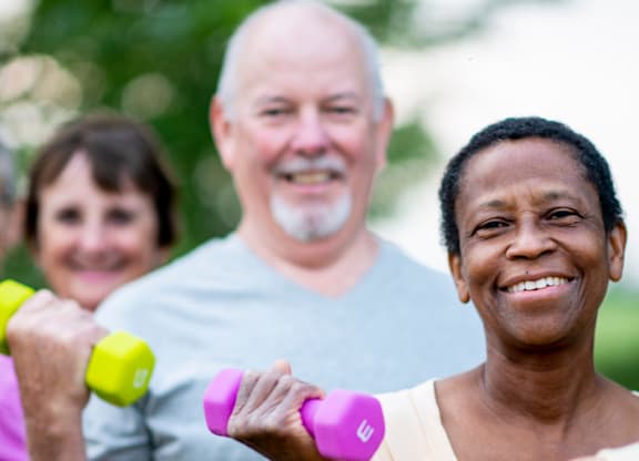 Three people exercising with dumbbells in a park.