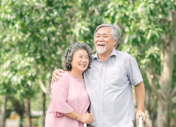 An elderly couple is standing in a park with the woman holding a walker.