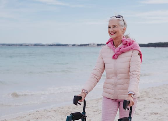 A woman in a pink jacket and pants is pushing a walker on a beach.