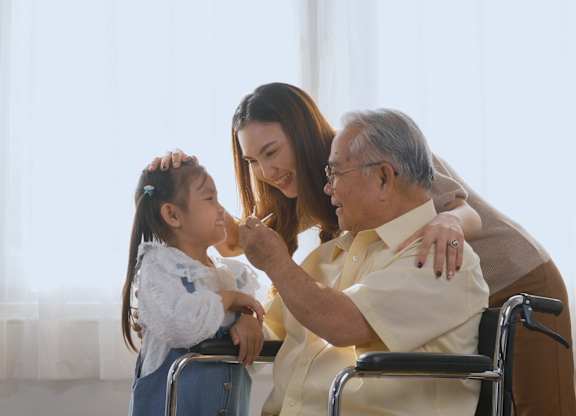 A woman is giving a young girl a haircut while an elderly man sits in a wheelchair nearby.