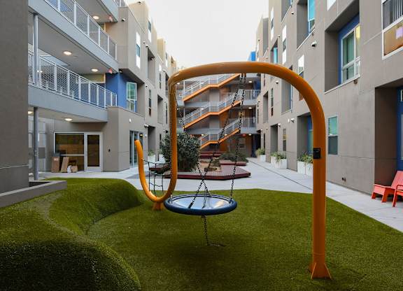 A playground with a swing set in the middle of a grassy area in front of apartment buildings.