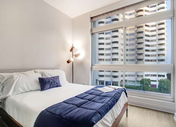 A bedroom with a bed, a window with a view of a building, and a wall lamp.at Wilshire Margot, Los Angeles, 90024 
