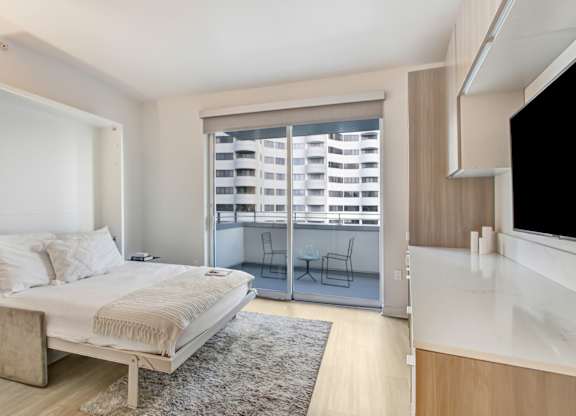 A bedroom with a bed, a chair, and a window with blinds.at Wilshire Margot, Los Angeles 