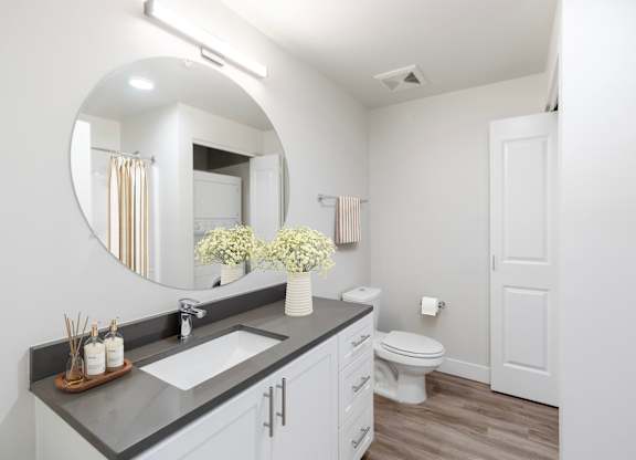A bathroom with a round mirror, sink, toilet, and wooden floors.
