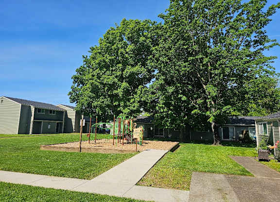 A playground with a slide and swings is surrounded by a grassy area and trees.