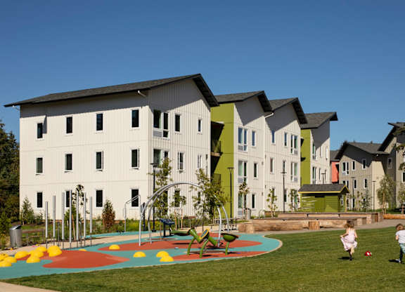 A playground with a colorful play structure is surrounded by a grassy area in front of a white building with a green roof.