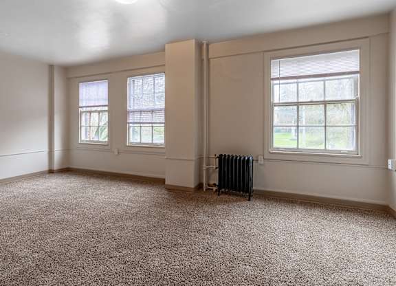 A room with a carpeted floor, two windows, and a radiator.