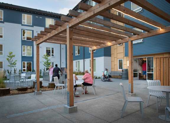 A wooden pergola is over a patio with people sitting and a motorcycle parked outside a building.