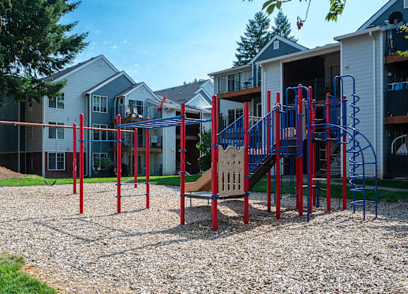 A playground with red and blue equipment in front of apartment buildings.