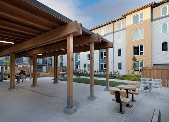 A wooden pergola is over a concrete patio with benches and picnic tables.