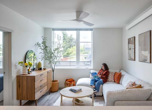 A woman is sitting on a couch in a living room with a bed, a table, and a plant.