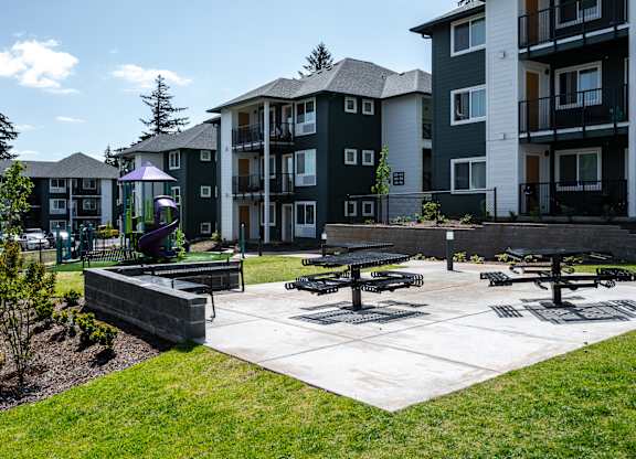 A park area with picnic tables and benches in front of apartment buildings.
