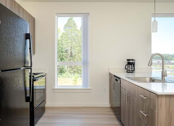 A black fridge stands in a kitchen with a window showing trees outside.