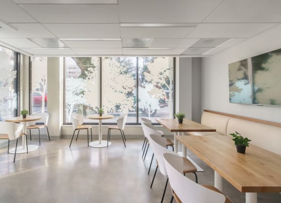 a room with wooden tables and white chairs with large windows in the background