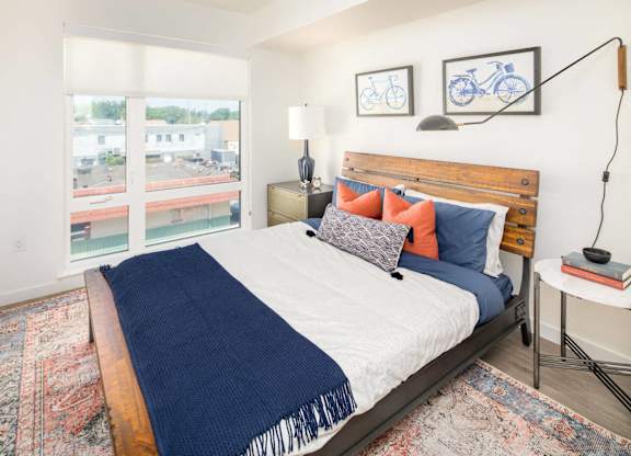 A bedroom with a bed, a nightstand, and a window overlooking a parking lot.