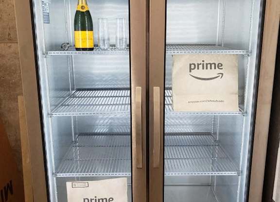 Two refrigerators, one with a bottle of champagne and one with a Prime bag.