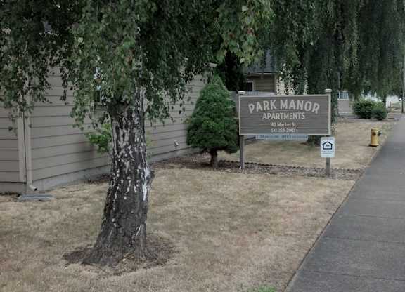A sign for Park Manor Apartments is on a post in front of a tree.