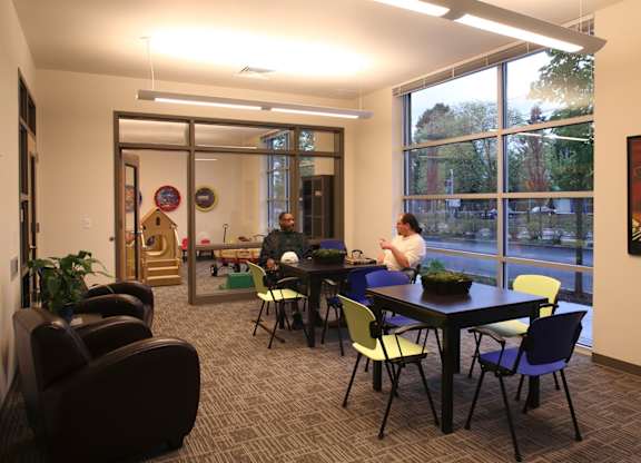 A conference room with a long table and chairs, a person standing at the head of the table, and a view of trees outside the window.