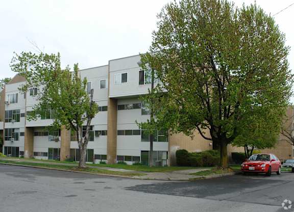A red car is parked on the street in front of a white apartment building.
