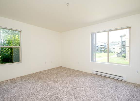 A room with a carpet floor, a window with a view of a building and trees outside, and a wall-mounted air conditioner.
