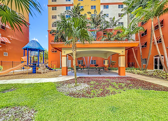A playground area with a slide and a gazebo is surrounded by palm trees and a building.