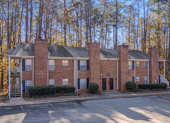 A red brick building with a black roof and chimneys is surrounded by trees.