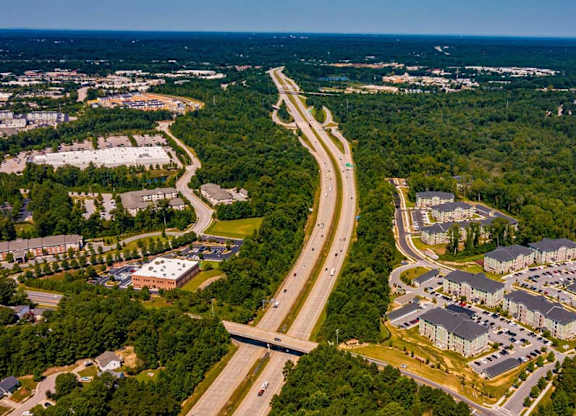 A highway curves through a wooded area with apartment buildings on one side and a parking lot on the other at Foxwood Apartments, North Carolina