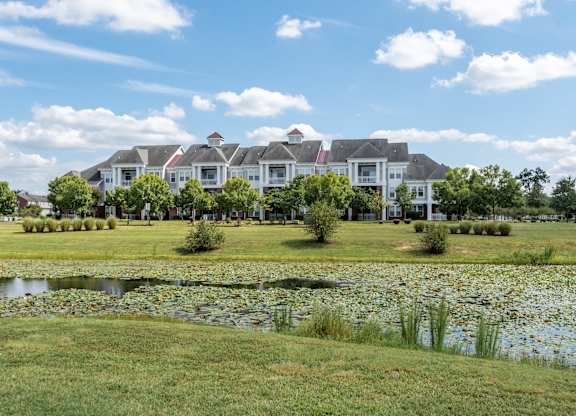 A large white house with a red roof is surrounded by a grassy lawn and a pond with lily pads.