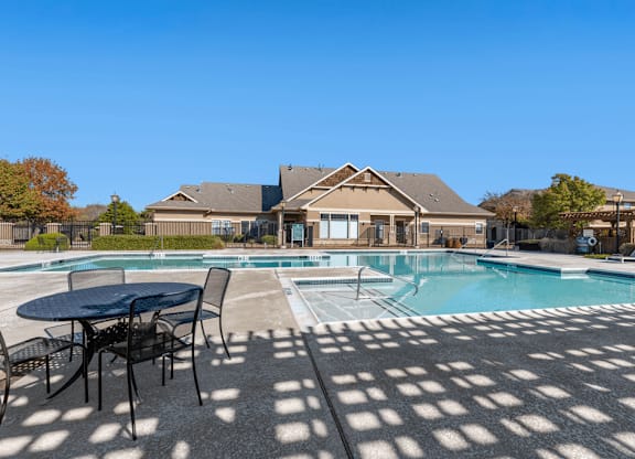 Swimming pool with large tanning deck, lounge chairs, round table and chairs with clubhouse exterior in background and native landscape