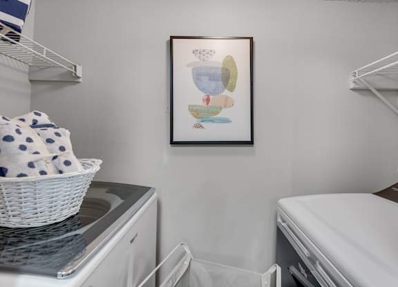 A laundry room with a washer and dryer, a basket on top of the washer, and a framed picture on the wall.