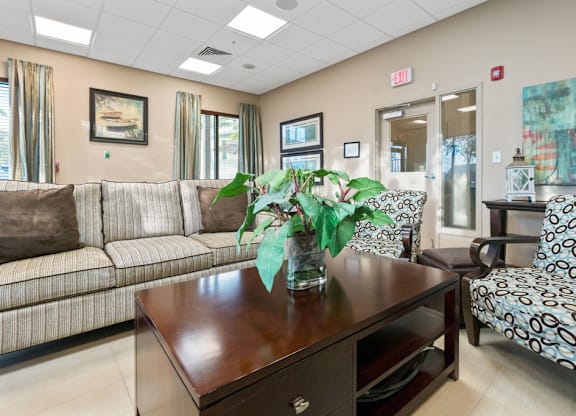 A room with a brown sofa, a patterned chair, and a wooden table with a vase of flowers on it.