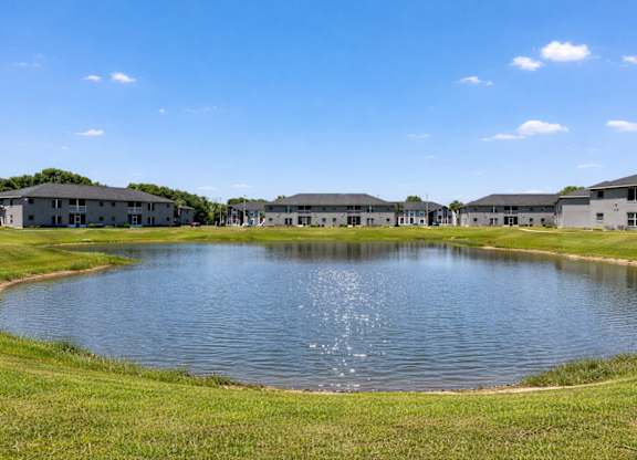 A large pond in the middle of a grassy field with buildings in the background.