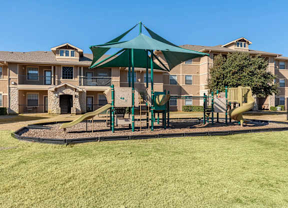 A playground with a green canopy and a yellow slide is in front of apartment buildings.