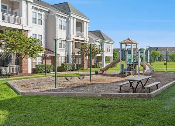 A playground with a swing set and picnic table in front of apartment buildings.