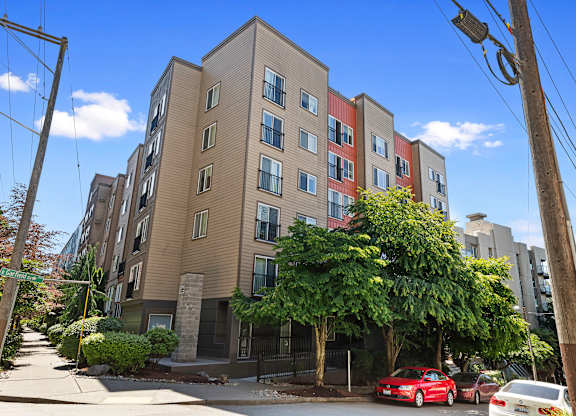 Kitty Corner View of apartment building on a street corner with cars parked, blue skies, and large green trees at Illumina Apartment Homes, Seattle