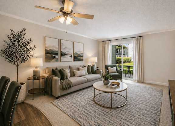 A living room with a grey sofa, a round table, a ceiling fan, and a painting on the wall.