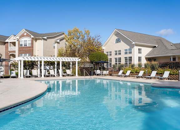 A large swimming pool in front of a building with a white pergola and lounge chairs. at Village on the Lake Apartments, Apartments, Spring Lake, NC