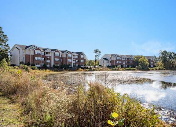 A row of apartment buildings is in the background of a body of water with a yellow flower in the foreground. at Village on the Lake Apartments, Spring Lake, North Carolina