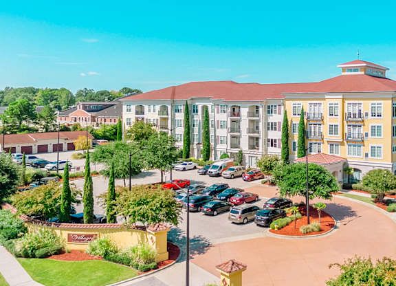 A large building with a red roof and a parking lot in front at The Villagio Apartments, Apartments, Fayetteville, NC