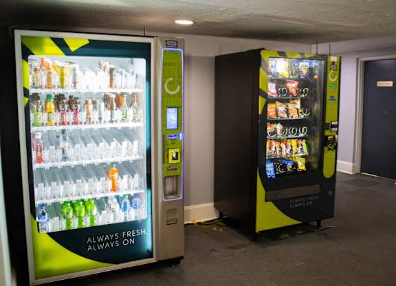 Two vending machines, one with a green and yellow color scheme, the other with a black and yellow color scheme, are in a room with a grey floor and a white ceiling.